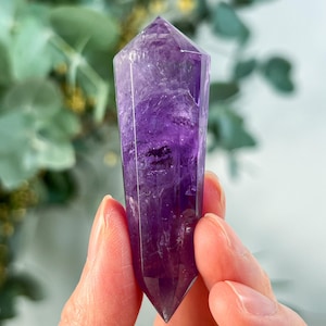 May include: A polished amethyst crystal point, showcasing a deep purple hue with lighter internal patterns. The crystal is held up against a blurred green and white background, highlighting its faceted shape and natural beauty. A gemstone for spiritual practices.