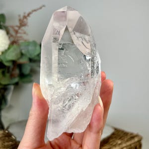 Puede incluir: Una punta de cristal de cuarzo transparente con una parte superior facetada y una base rugosa y texturizada. El cristal se sostiene en una mano, mostrando su transparencia y formaciones naturales. El fondo incluye vegetación y una cesta de mimbre.
