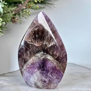 Könnte beinhalten: Ein polierter Amethystkristall in Tropfenform. Der Kristall zeigt Farbtöne von tiefem Lila, Lavendel und Braun, mit sichtbaren inneren Mustern und Einschlüssen. Er steht auf einer weißen Oberfläche mit goldenem Rand, mit einem grünen Zweig im Hintergrund.