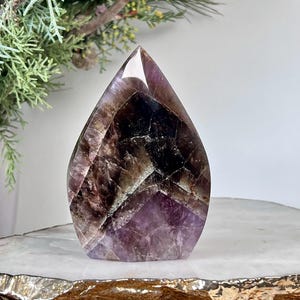 Könnte beinhalten: Ein polierter Amethystkristall in Tropfenform. Der Kristall weist Schattierungen von Lila, Braun und Weiß auf, mit sichtbaren inneren Mustern. Er steht auf einer weißen und goldfarbenen Oberfläche, mit Grün im Hintergrund.