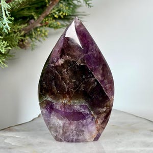 Könnte beinhalten: Ein polierter Amethystkristall in Tropfenform. Der Kristall weist Farbtöne von tiefem Lila, Braun und Hauch von Grün auf. Der Stein wird auf einer weiß-goldfarbenen Oberfläche mit einer grünen Pflanze im Hintergrund präsentiert.