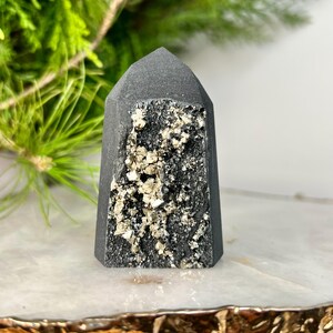 May include: A dark gray crystal tower with a flat top and a rough, textured surface. The center of the tower features a cluster of sparkling, gold-colored pyrite crystals. The crystal is displayed on a marble surface.