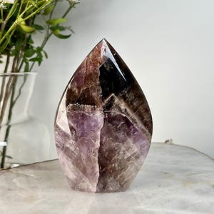Könnte beinhalten: Ein polierter Amethyst-Kristallpunkt in Tropfenform. Der Kristall zeigt eine Mischung aus violetten, braunen und klaren Farbtönen mit sichtbaren inneren Mustern. Der Sockel ist hell und der Kristall steht auf einer weißen Oberfläche.