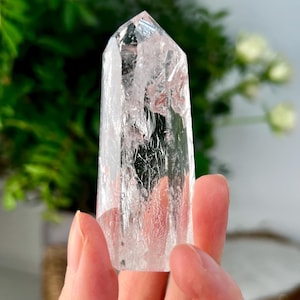 Puede incluir: Una punta de cristal de cuarzo transparente, sostenida frente a un fondo borroso verde y blanco. El cristal es transparente con fracturas internas y una parte superior puntiaguda. El cristal mide aproximadamente 10 cm de alto.