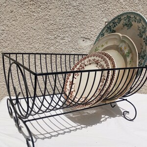 May include: A black wire dish rack with a curved, open-frame design and scroll-style feet. The rack holds three ceramic plates, including one with a brown floral pattern, one with a green floral design, and one with a blue floral border.