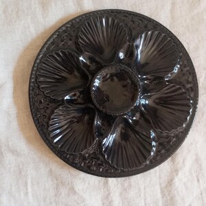 May include: A round, dark brown ceramic oyster plate featuring six shell-shaped indentations arranged in a circular pattern around a central circular well. The surface has a glossy finish with a textured, pebbled border along the outer edge.