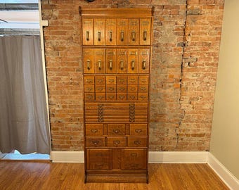 May include: A tall, antique wooden cabinet with numerous drawers and compartments. The cabinet is made of light brown wood and features brass handles. It stands against a brick wall and hardwood floor, suggesting a vintage or retro style.