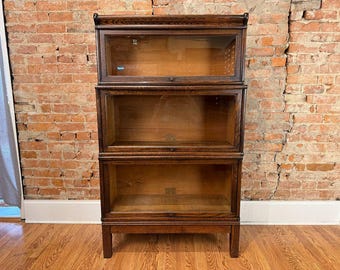 Op de afbeelding: Een drielaags boekenkast van donker hout met glazen planken. De kast in antieke stijl heeft een rechthoekige vorm en staat op korte poten. De achtergrond is voorzien van een bakstenen muur en een hardhouten vloer.