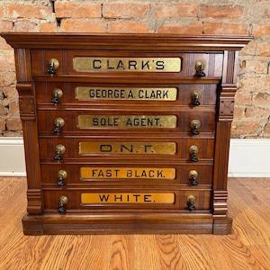 May include: A vintage wooden cabinet with six drawers, each labeled with gold lettering. The labels read "CLARK'S", "GEORGE A. CLARK", "SOLE AGENT", "O.N.T.", "FAST BLACK", and "WHITE". The cabinet has ornate hardware and is set against a brick wall.