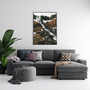 May include: A gray sectional sofa with a throw blanket and pillows in various patterns and colors. A framed photograph of a waterfall in a rocky landscape hangs above the sofa.