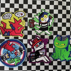 May include: A collection of colorful cartoon cat stickers on a black and white checkered background. The stickers feature various cat designs with speech bubbles, holding objects, and wearing 3D glasses. One sticker reads "BAD THING".