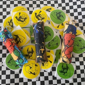 Op de afbeelding: Een assortiment kleurrijke sleutelhangers en stickers. De sleutelhangers tonen karakterkunst in rood, blauw en zwart. De stickers zijn rond met gele en groene achtergronden en zwarte karakterontwerpen. De items zijn gerangschikt op een zwart-wit geblokt oppervlak.