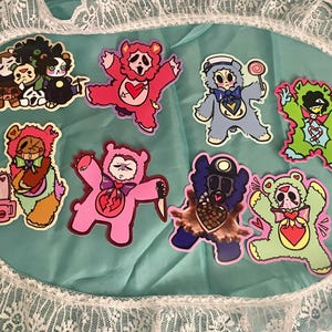 May include: A set of nine colorful cartoon bear stickers. The bears are wearing different outfits and have different expressions. Some of the bears are holding objects, such as a chainsaw, a lollipop, and a knife. The stickers are perfect for adding a touch of whimsy to any surface.