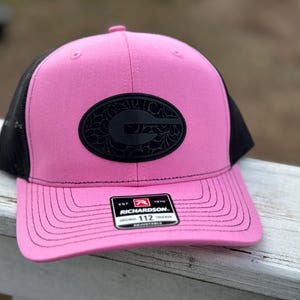 Peut inclure: Casquette de camionneur rose et noire avec un logo brodé noir. Le logo est une lettre stylisée "G" avec un motif floral. La casquette a un dos en maille noire et un devant rose avec une visière cousue noire. La casquette a une étiquette noire avec le texte "EST. 1970 RICHARDSON 112 ADJUSTABLE".