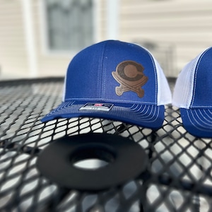 May include: Two blue and white trucker hats with a brown leather patch featuring a stylized "C" with crossed swords. The hats are on a black metal table.