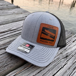 May include: A gray and black trucker hat with a brown leather patch that says "I Hate Pulling Out". The patch features a silhouette of a boat. The hat is made by Richardson 112.
