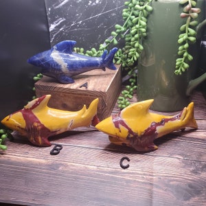 May include: Three carved stone shark figurines. One is blue with white veining, and the other two are yellow with red and brown veining. The sharks are displayed on a wooden surface with a green mug and greenery in the background.