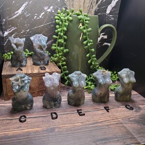 May include: Six green and white agate stone sculptures of female torsos, arranged in a row on a wooden surface. The sculptures are labeled A through F.