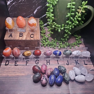 May include: A collection of polished gemstone eggs in various colors and patterns, including red, orange, brown, green, blue, white, and pink. The eggs are displayed on a wooden surface with letter labels A through L.