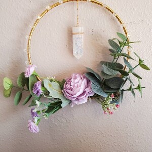 Puede incluir: Una corona de alambre dorado con un colgante de cristal transparente que cuelga en el centro. La corona está decorada con flores artificiales moradas y verdes.