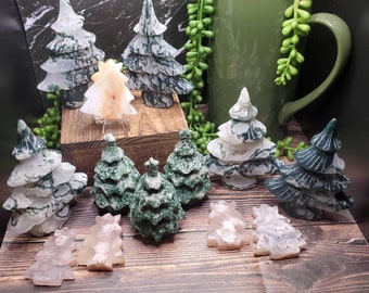 May include: A collection of green and white agate Christmas tree ornaments. The trees are carved from natural agate and have a variety of patterns and colors. Some of the trees are standing on a wooden surface, while others are lying on a table. There are also a few pink and white agate ornaments in the shape of Christmas trees.