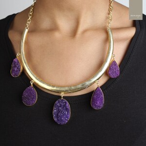 May include: A gold necklace with a thick, curved bar and five dangling purple amethyst stones. The stones are all different shapes and sizes, and they have a rough, textured surface.