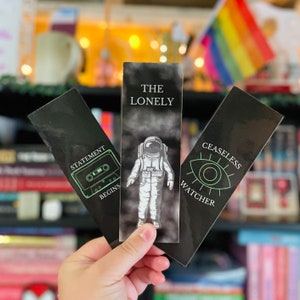 May include: Three black bookmarks with white and neon green graphics. One bookmark reads "STATEMENT BEGINS" with a cassette tape graphic. Another says "THE LONELY" with an astronaut. The third says "CEASELESS WATCHER" with an eye graphic.