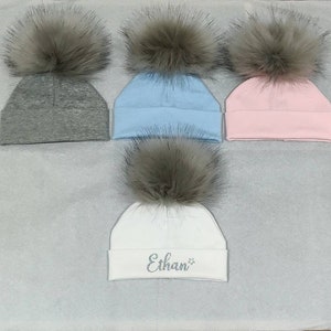 May include: Four knit hats with faux fur pom poms. Three hats are solid colors: gray, light blue, and pink. The fourth hat is white with the name "Ethan" embroidered in silver glitter.