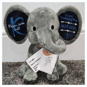 Op de afbeelding: Een grijze pluche olifant met zwarte details. De oren van de olifant hebben donkerblauwe patches met witte tekst en blauwe glitter. Op &eacute;&eacute;n oor staat "Reggie James Scott" en op het andere "On your Christening Day 01.08.2021". Een label leest "NUGS & KISSES".