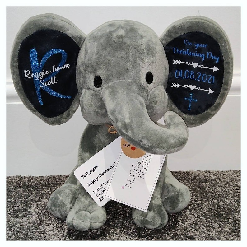 Op de afbeelding: Een grijze pluche olifant met zwarte details. De oren van de olifant hebben donkerblauwe patches met witte tekst en blauwe glitter. Op &eacute;&eacute;n oor staat "Reggie James Scott" en op het andere "On your Christening Day 01.08.2021". Een label leest "NUGS & KISSES".