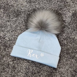 May include: A light blue knitted baby hat with a white faux fur pom pom and the name "Rex" embroidered on the front in white with a white star.