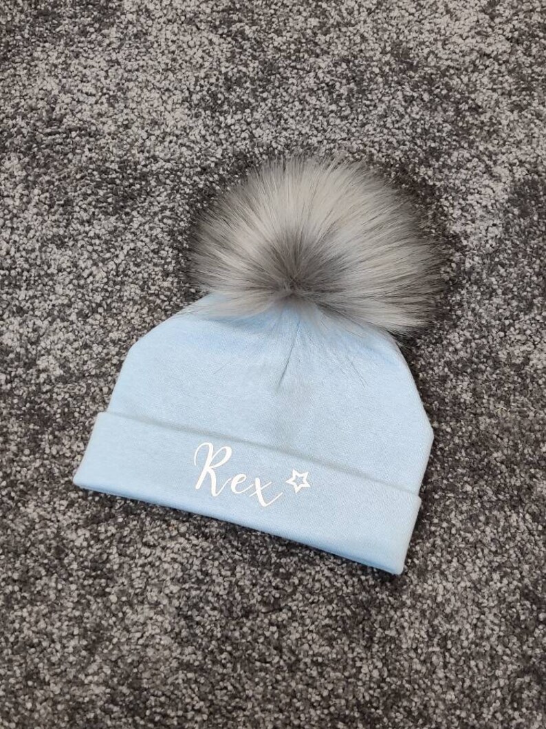 May include: A light blue knitted baby hat with a white faux fur pom pom and the name "Rex" embroidered on the front in white with a white star.
