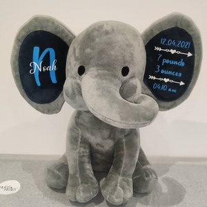 May include: Gray plush elephant stuffed animal with blue embroidered details. The elephant's ears have black fabric with the name "Noah" and birth details: "12.04.2021, 7 pounds 3 ounces, 04:10 am" embroidered in blue.