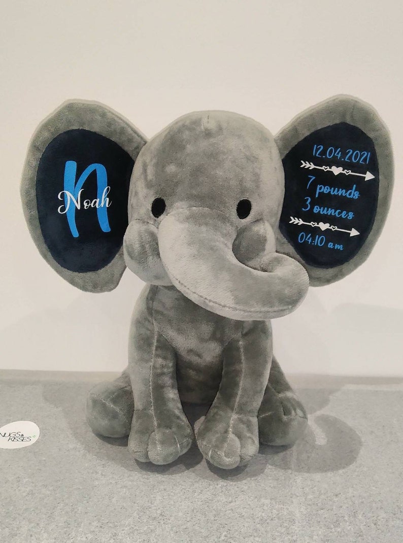 May include: Gray plush elephant stuffed animal with blue embroidered details. The elephant's ears have black fabric with the name "Noah" and birth details: "12.04.2021, 7 pounds 3 ounces, 04:10 am" embroidered in blue.