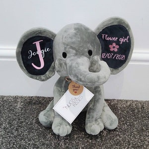 Op de afbeelding: Een grijze pluche olifant met zwarte ooraccenten. Het linkeroor heeft de naam "Jorgie" in roze script, en het rechteroor zegt "Flower girl" met een roze bloem en de datum 12/07/2021. Een label leest "Nugs & Kisses" en "Thank you!"