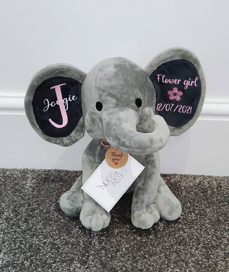 Op de afbeelding: Een grijze pluche olifant met zwarte ooraccenten. Het linkeroor heeft de naam "Jorgie" in roze script, en het rechteroor zegt "Flower girl" met een roze bloem en de datum 12/07/2021. Een label leest "Nugs & Kisses" en "Thank you!"