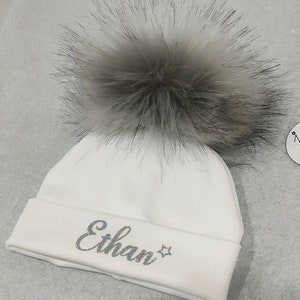 May include: White knitted baby beanie with a fluffy gray pom pom and the name "Ethan" embroidered in silver glitter.