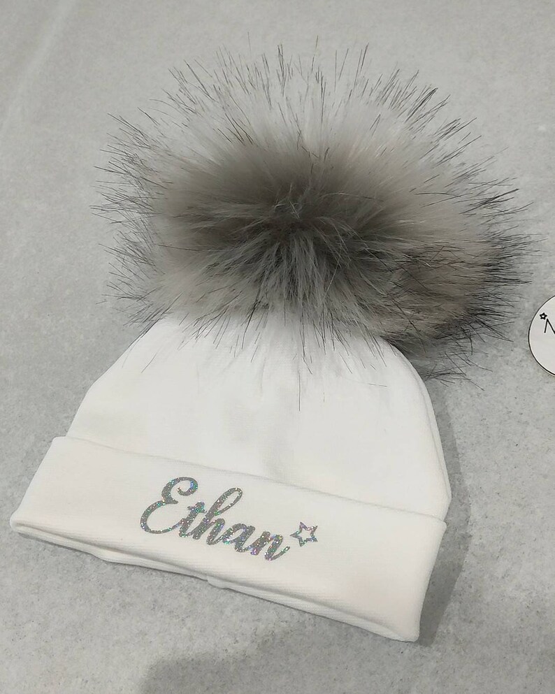 May include: White knitted baby beanie with a fluffy gray pom pom and the name "Ethan" embroidered in silver glitter.