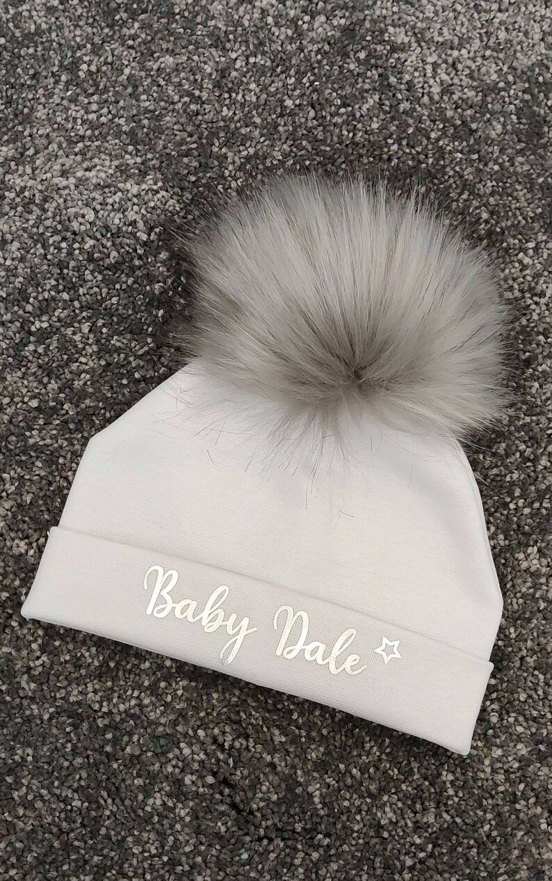 May include: White baby hat with a fluffy pom pom and the text "Baby Dale" embroidered in silver.