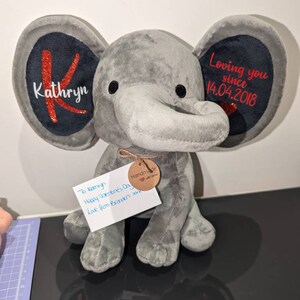 Op de afbeelding: Een grijze pluche olifant met grote oren. Het linkeroor heeft de naam "Kathryn" en een rode "K". Het rechteroor leest "Loving you since 14.04.2018". Een label leest "Handmade with love".