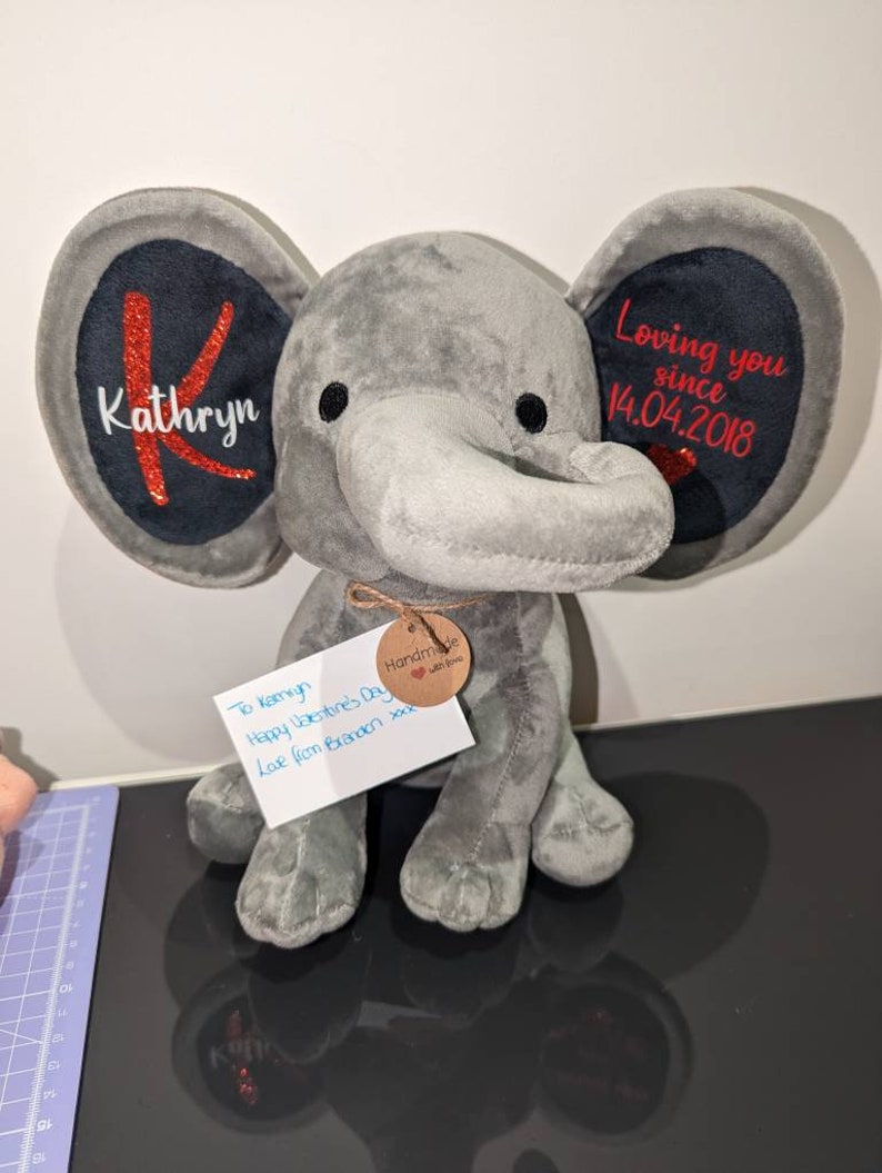 Op de afbeelding: Een grijze pluche olifant met grote oren. Het linkeroor heeft de naam "Kathryn" en een rode "K". Het rechteroor leest "Loving you since 14.04.2018". Een label leest "Handmade with love".