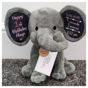 Op de afbeelding: Een grijze pluche olifant met geborduurde details. Het linkeroor leest "Happy 1st Birthday Alaya". Het rechteroor toont geboortegegevens: 15 juni 2020, 3,6 kg, en 22:22 uur. Een label leest "Handmade with love, Nugs & Kisses."