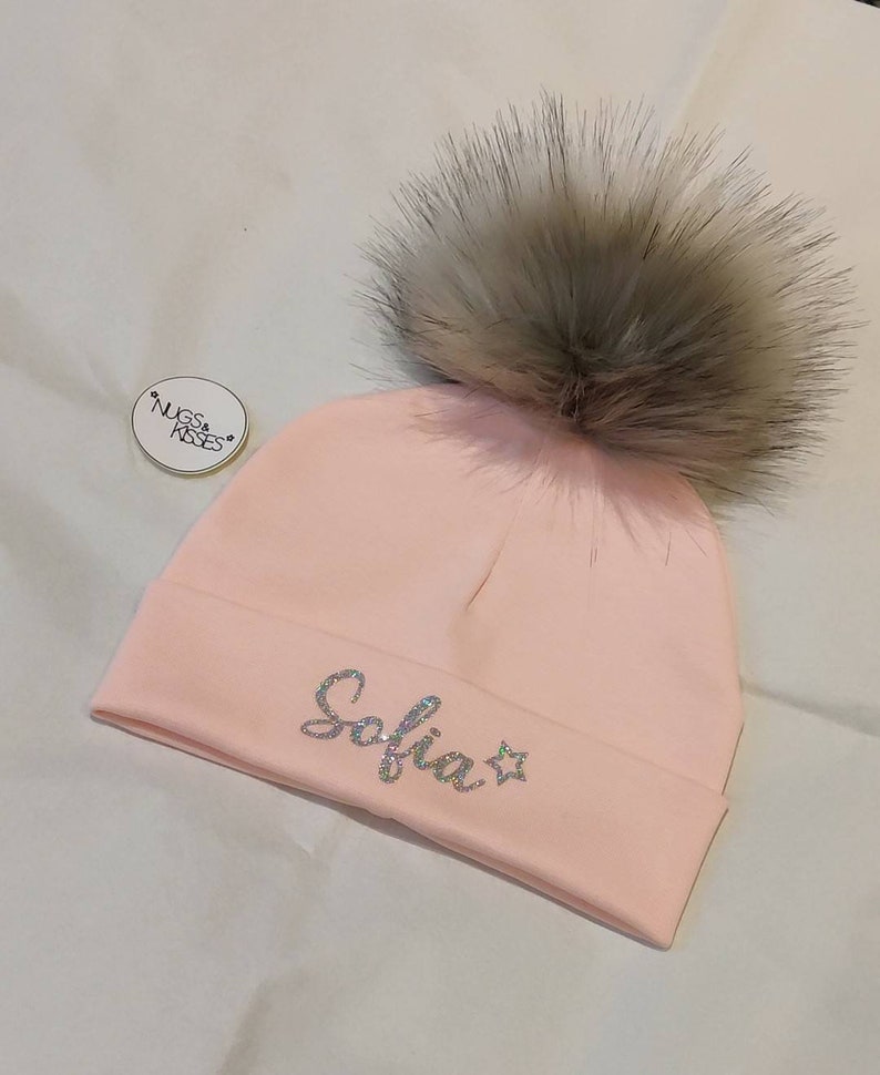 May include: Pink knit beanie with a fluffy gray pom pom and the name "Sofia" in sparkly silver letters with a star.