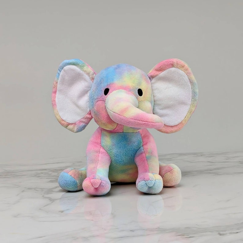 May include: A plush tie-dye elephant toy with a pink, blue, and yellow color scheme. The elephant has white ears and a long, pink trunk.
