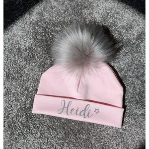 May include: Pink knitted baby hat with a fluffy white pom pom and the name "Heidi" in sparkly silver lettering.