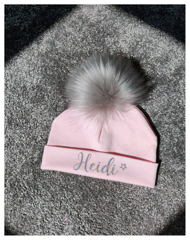 May include: Pink knitted baby hat with a fluffy white pom pom and the name "Heidi" in sparkly silver lettering.