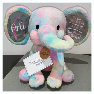 Op de afbeelding: Een pluche olifant in pastel regenboogkleuren. De olifant heeft grote oren met de tekst "Arli" en "On our Special Day 13.11.2021 Love Mummy & Daddy xx". Een label leest "NUGS & KISSES" en "Hand made with love".