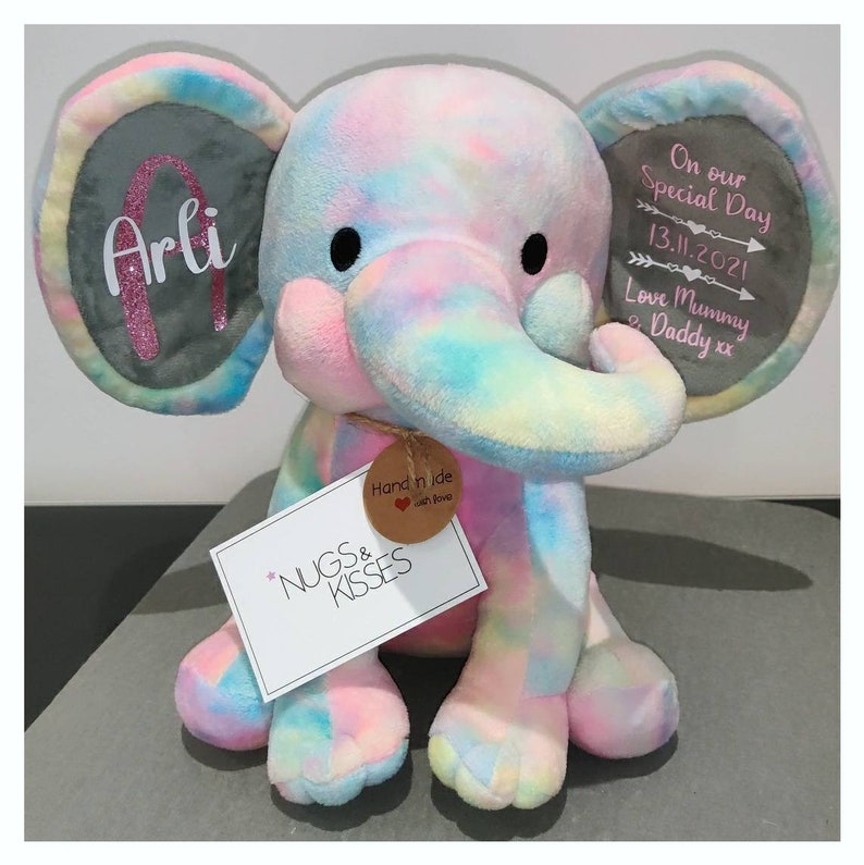 Op de afbeelding: Een pluche olifant in pastel regenboogkleuren. De olifant heeft grote oren met de tekst "Arli" en "On our Special Day 13.11.2021 Love Mummy & Daddy xx". Een label leest "NUGS & KISSES" en "Hand made with love".