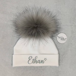 May include: White baby beanie with a fluffy gray pom pom and the name "Ethan" embroidered in silver glitter on the front. The beanie has a small tag attached with the text "NUGS & KISSES".