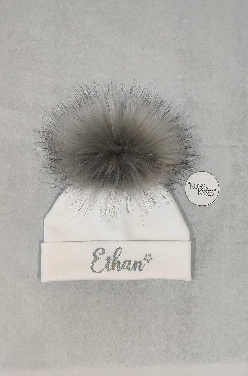 May include: White baby beanie with a fluffy gray pom pom and the name "Ethan" embroidered in silver glitter on the front. The beanie has a small tag attached with the text "NUGS & KISSES".